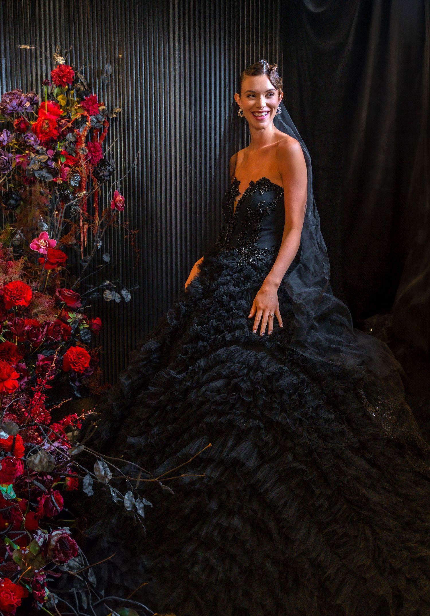 A person in a black ruffled ball gown poses beside red floral arrangements.