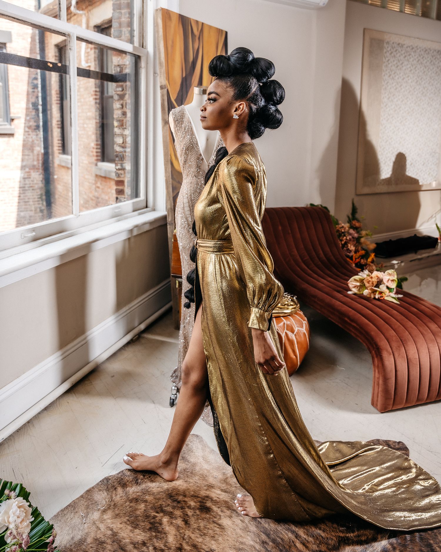Side profile of model in flowing gold dress posing by window with vintage decor.