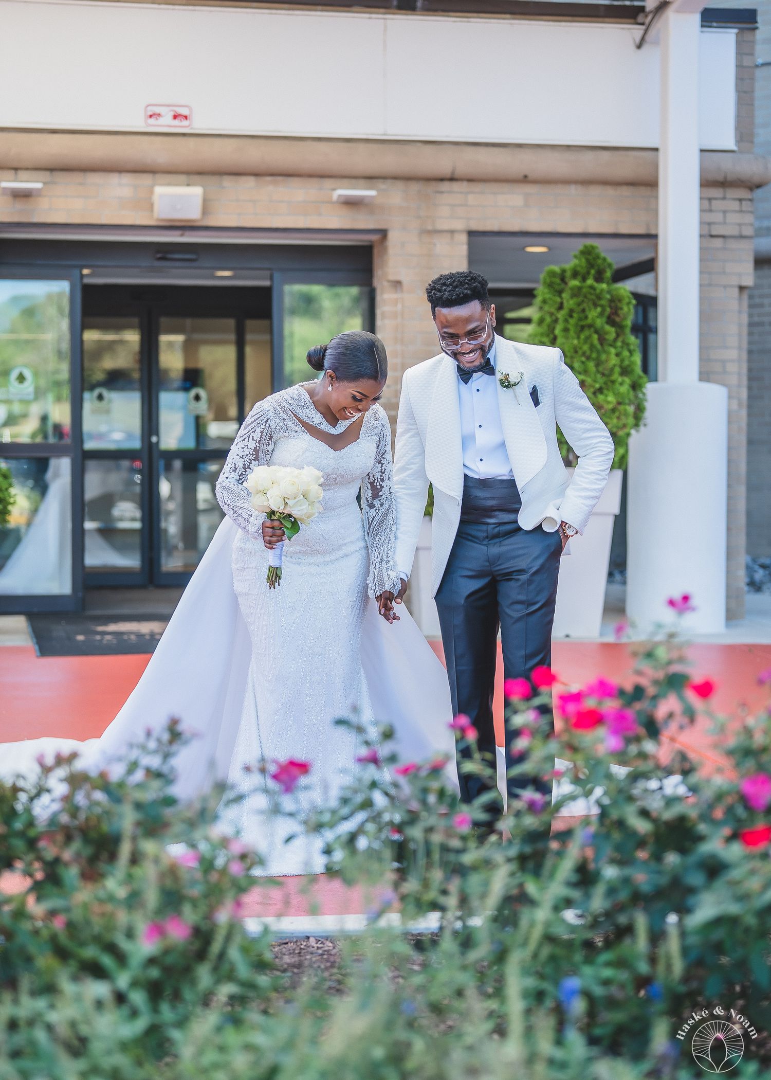 A joyful romantic moment between newlyweds in formal attire posing by a pink rose garden at their wedding.