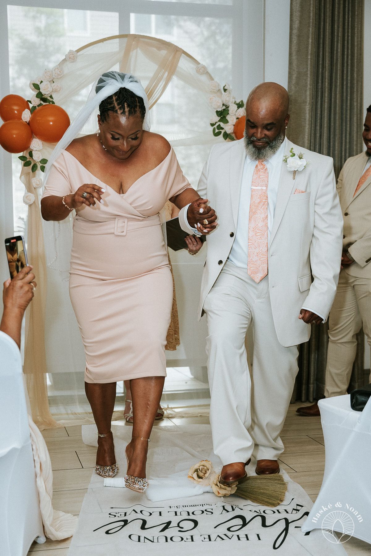 Wedding couple walks down aisle under decorated arch with orange balloons and greenery.