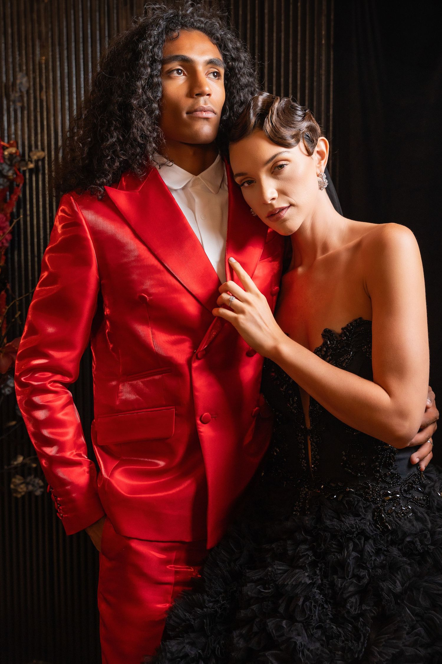 A glamorous couple poses intimately against a dark wall with red floral arrangements.
