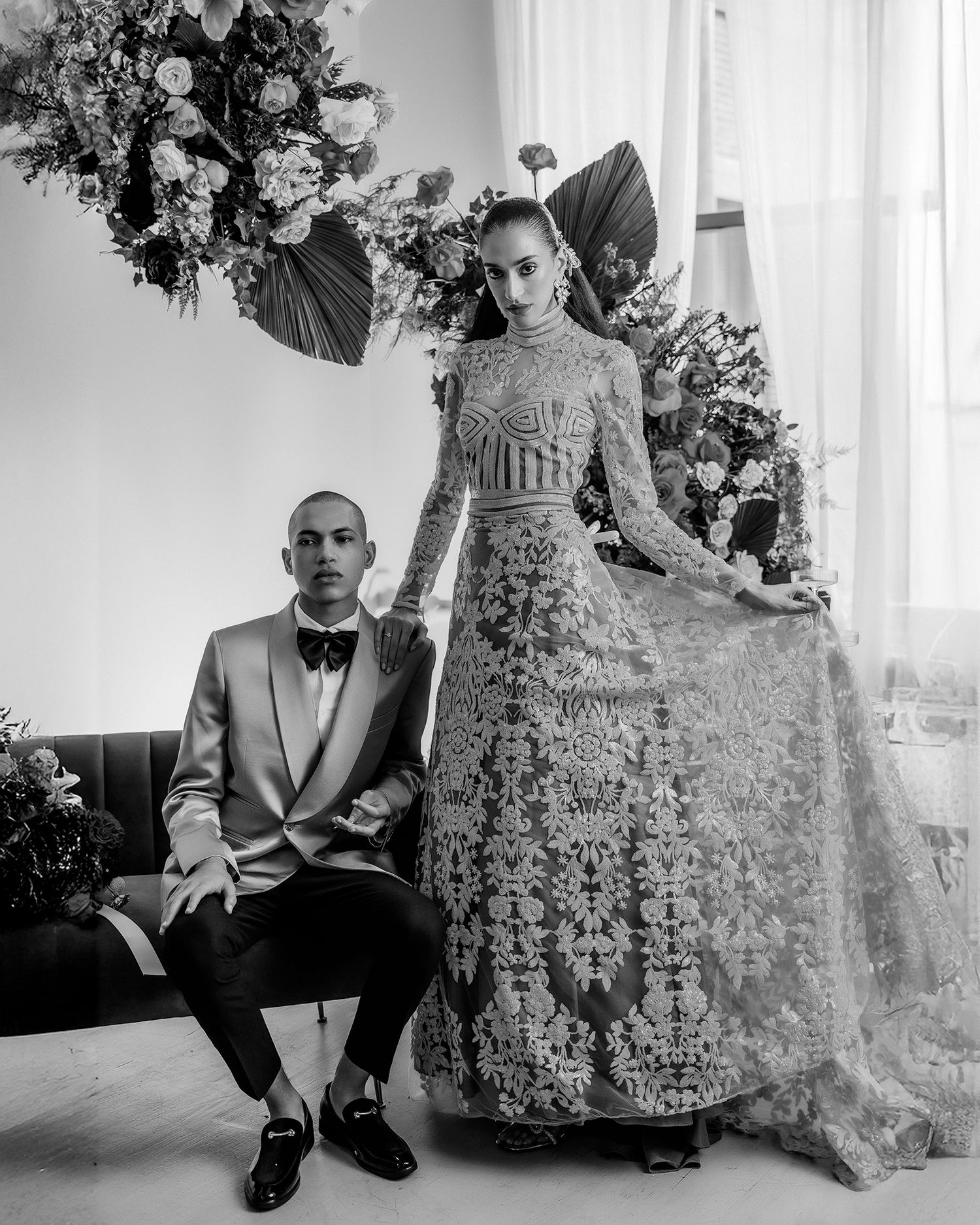 A glamorous couple poses in formal attire against a floral backdrop with dark red palm fans.