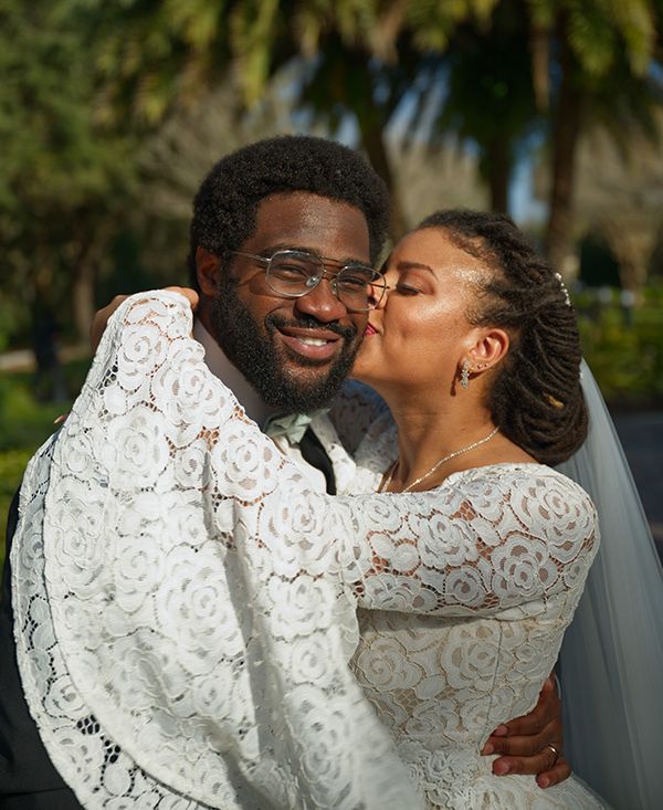 A joyful wedding couple in matching white lace outfits share an intimate moment under palm trees.