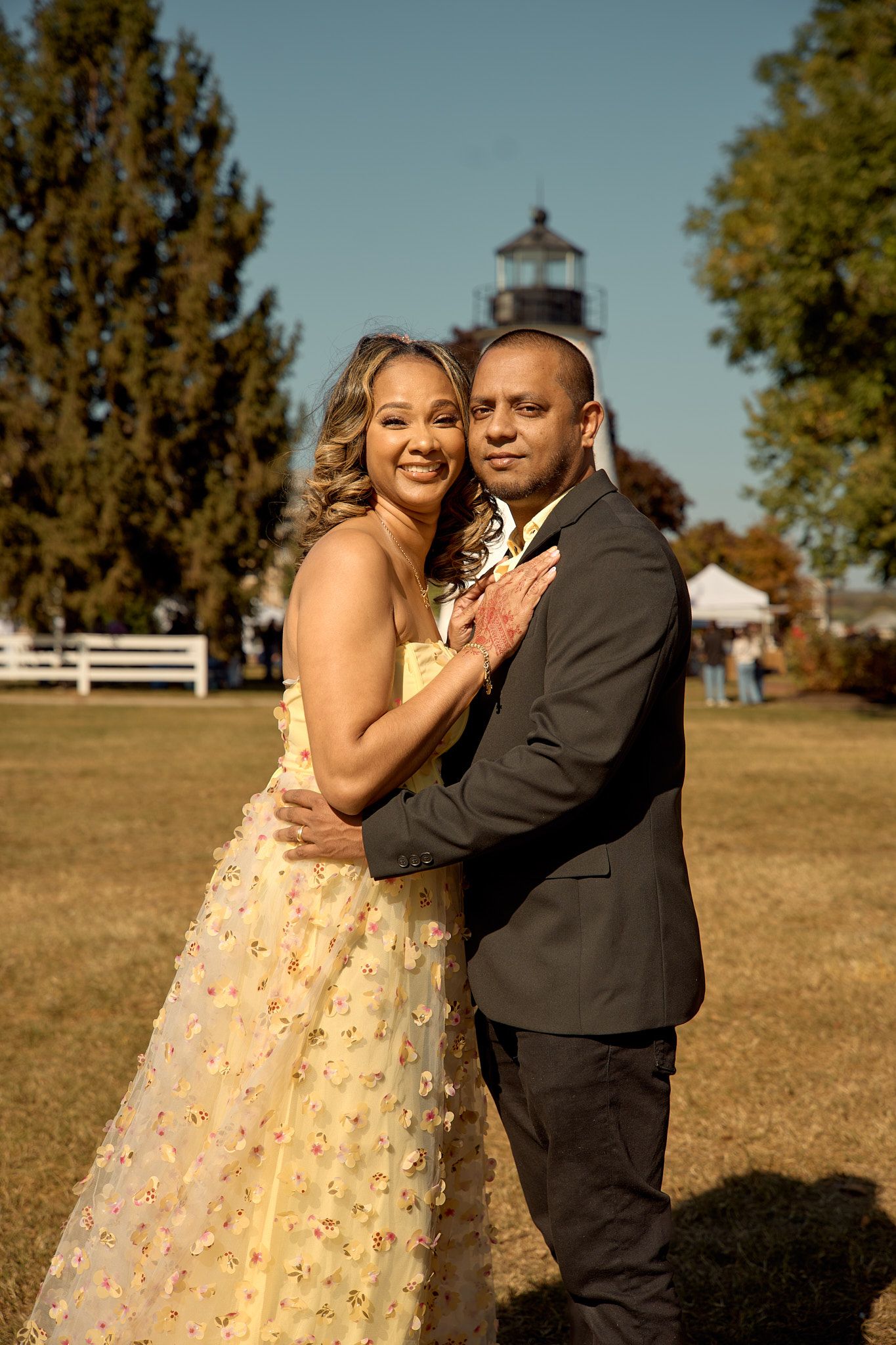 A couple in formal attire poses in front of a lighthouse on a sunny autumn day.