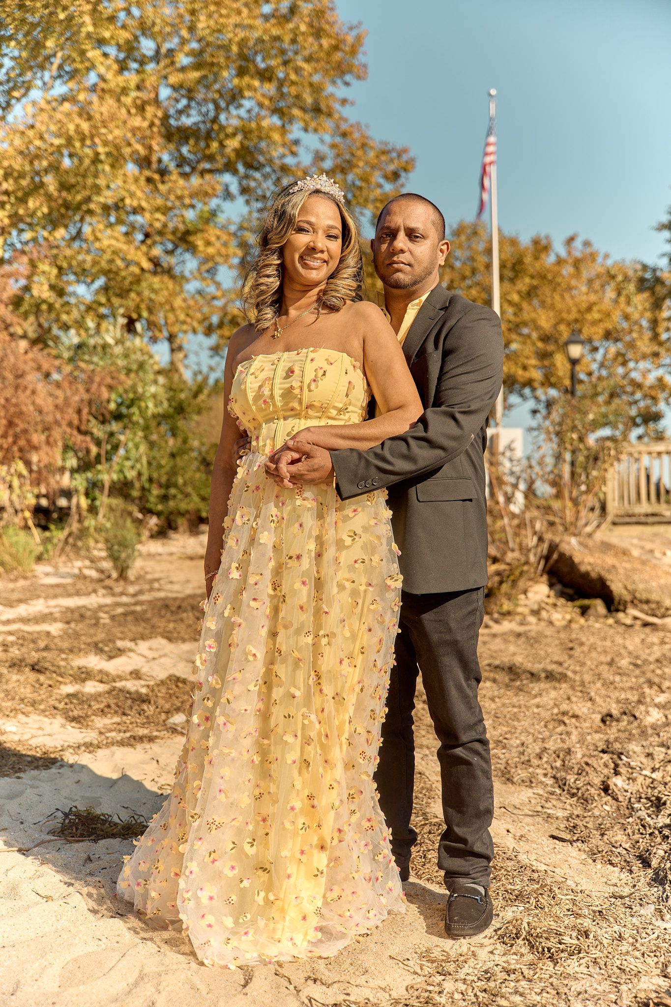 A couple poses together outdoors with an American flag and fall scenery in the background.