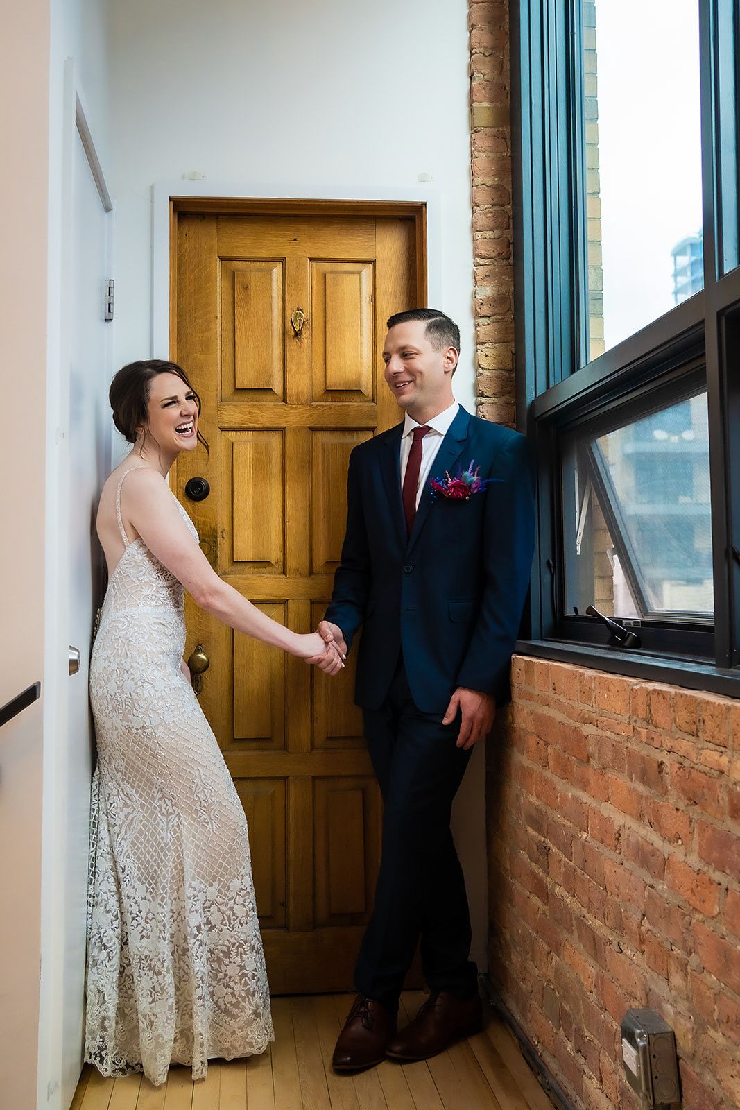 A couple shares a candid moment in wedding attire by a wooden door and brick wall.