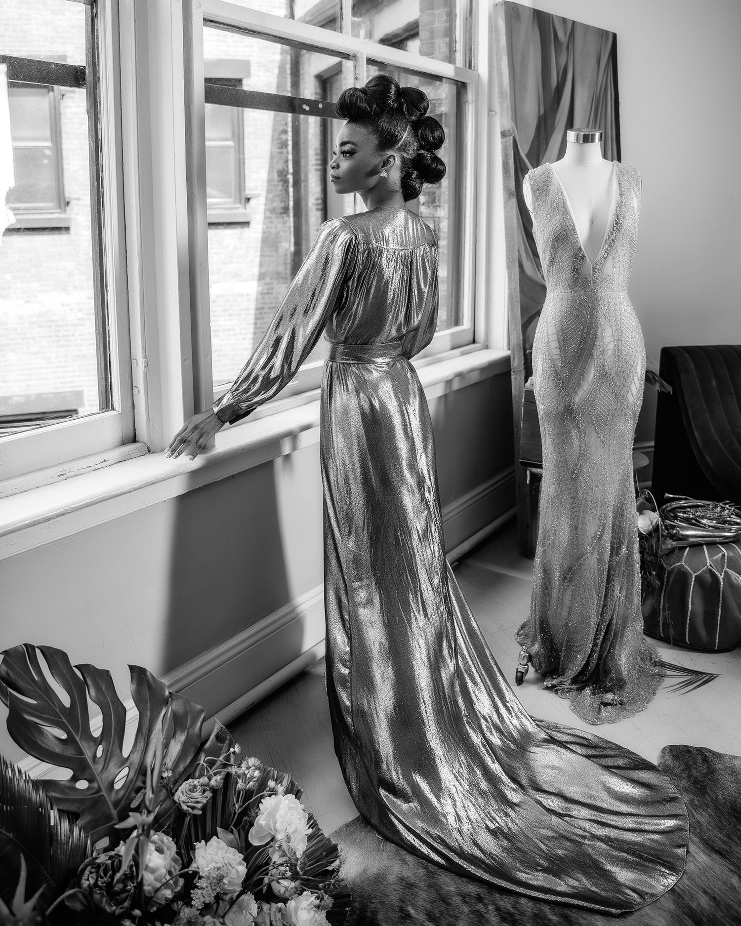 Elegant black and white photo of someone in a flowing evening gown next to a dress form by a window.