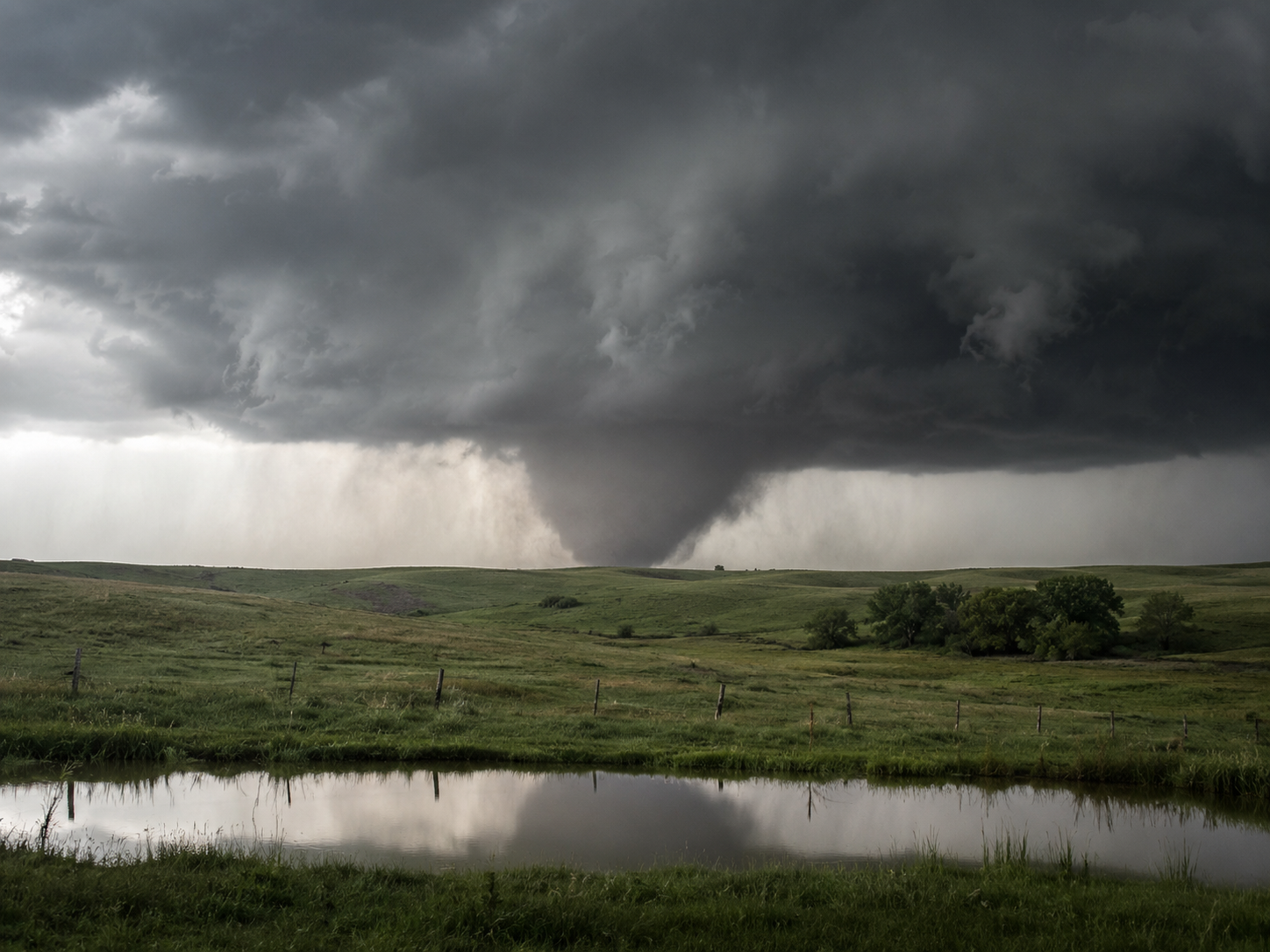 A massive tornado touches down over green rolling plains with a reflective pond in the foreground under dark stormy skies.