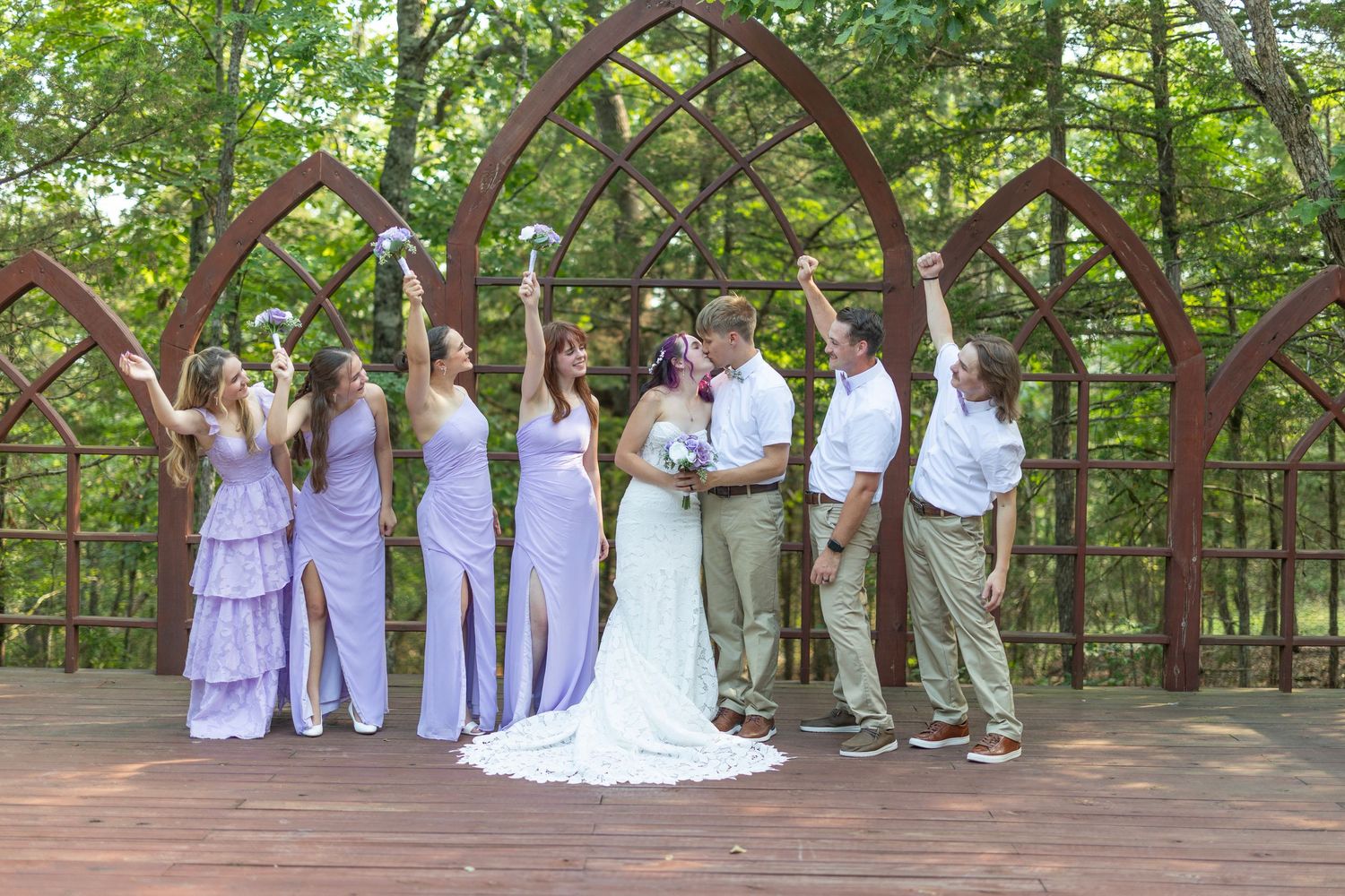 Wedding party celebrates on wooden deck with Gothic arches, bridesmaids in lavender dresses raising bouquets beside bride and groom.