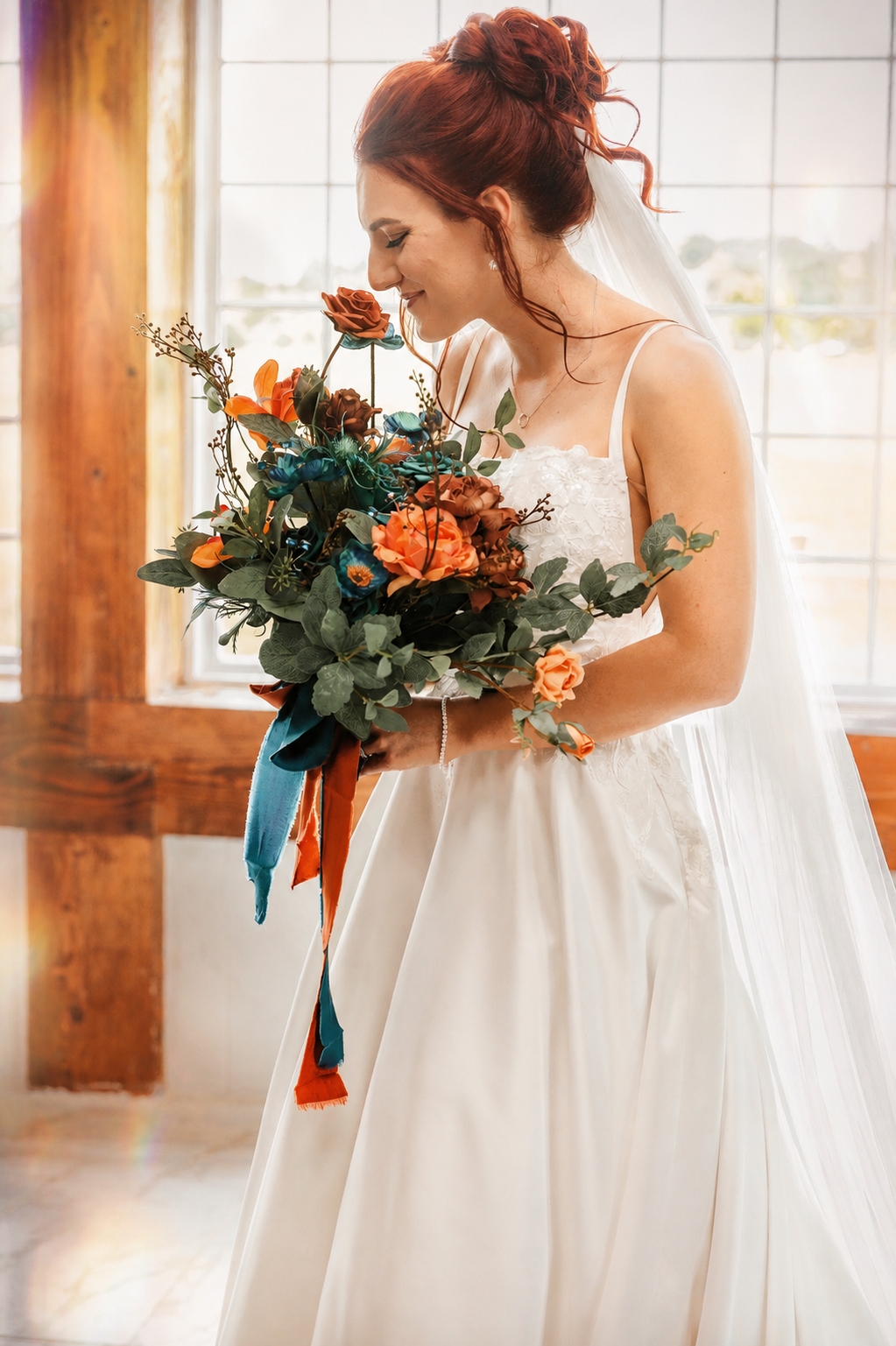 Smiling redhead bride in white spaghetti-strap gown holds a lush orange and teal bouquet with flowing ribbons in soft light.