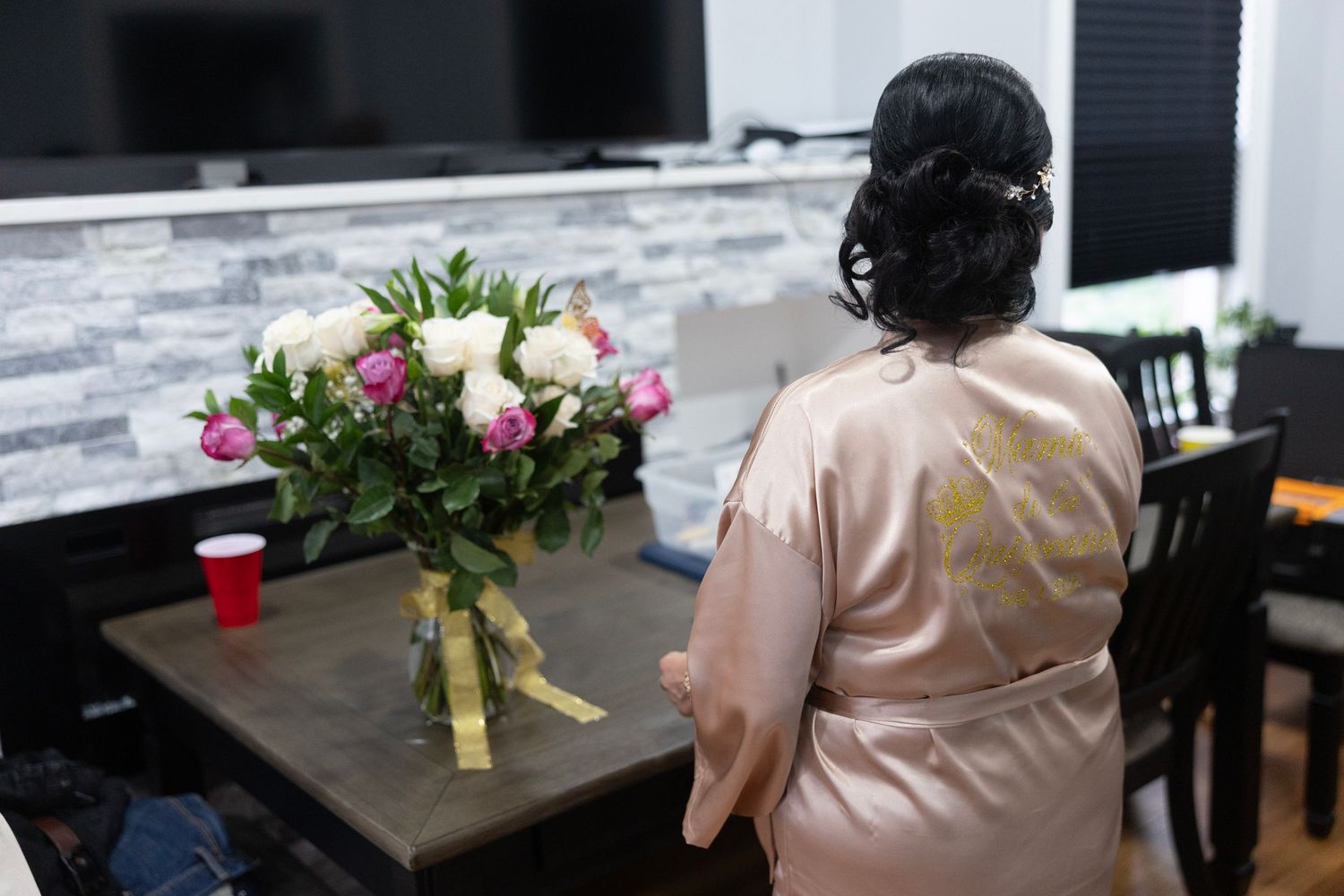 Woman in satin robe with updo hairstyle sits near a bouquet of pink and white roses at a wedding preparation.