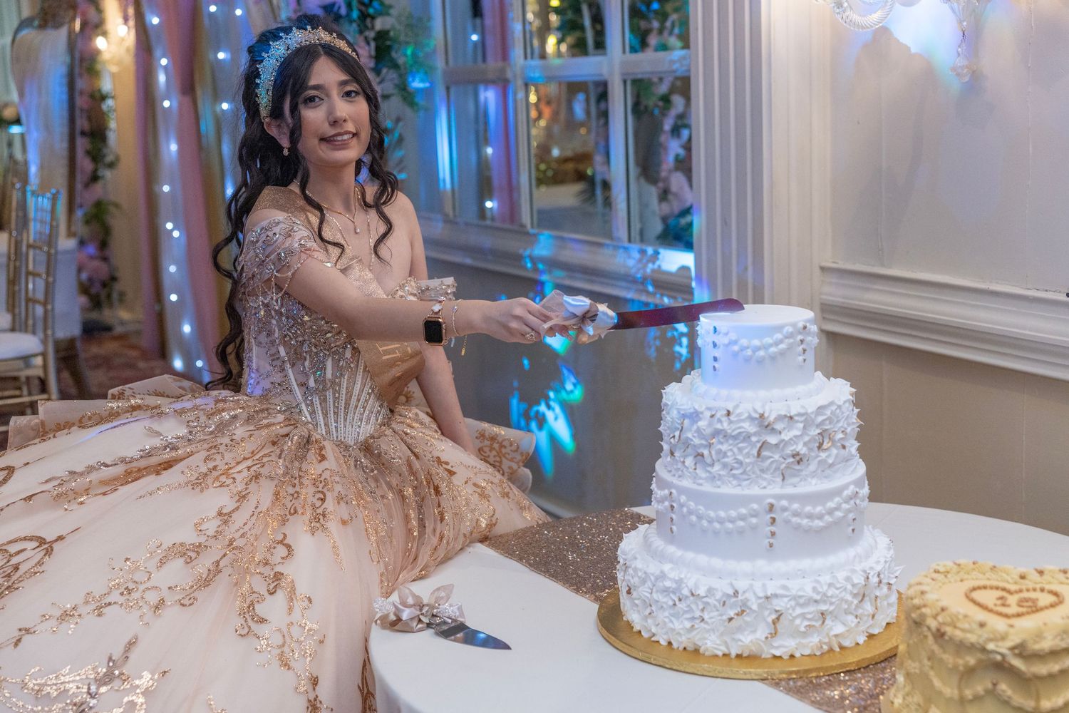 Young woman in elegant gold gown cutting a white tiered cake at a quinceañera celebration.
