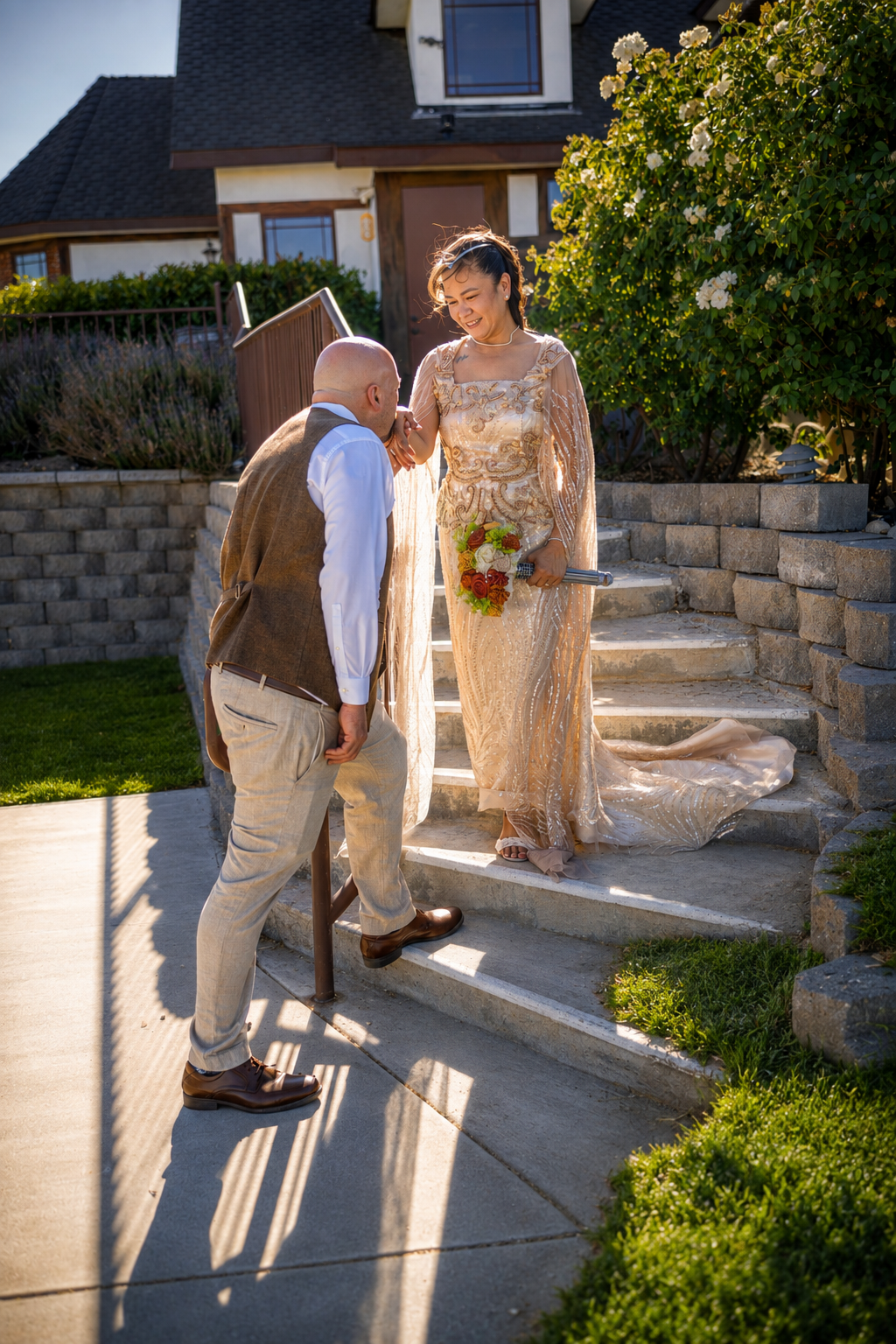 Groom kisses bride's hand on sunlit garden steps, she wears a gold lace gown holding a bouquet.