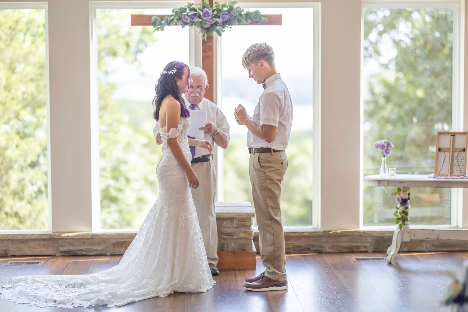 Bride and groom exchanging vows with officiant at elegant indoor wedding ceremony near large windows.