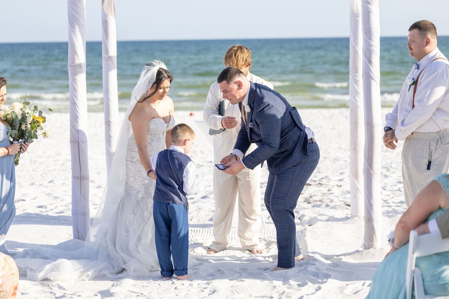 Bride and groom exchange rings with a young boy on a sunny beach wedding ceremony with white draping.
