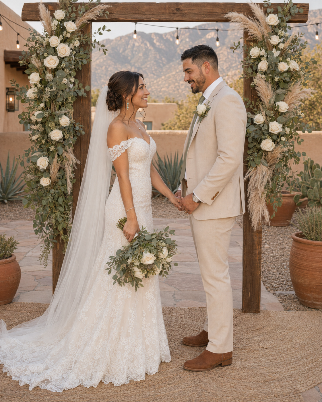 Bride and groom holding hands at outdoor desert wedding ceremony under wooden arch with white roses and greenery.