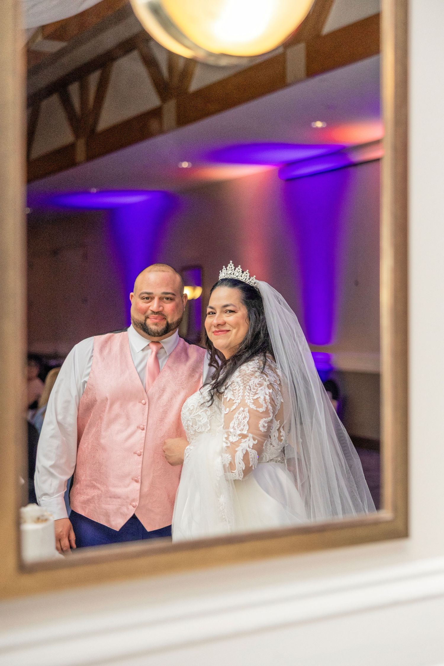 Bride and groom smiling in mirror reflection at wedding reception with purple uplighting in elegant venue.