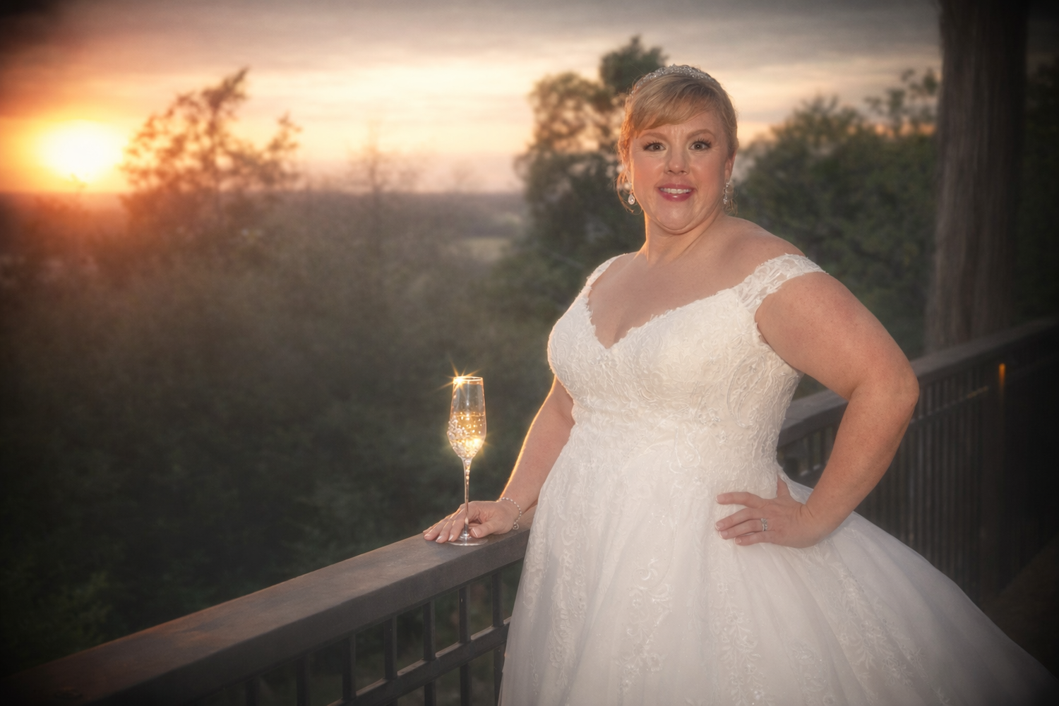 Bride in white gown holding champagne flute on balcony at sunset with scenic tree-lined backdrop.