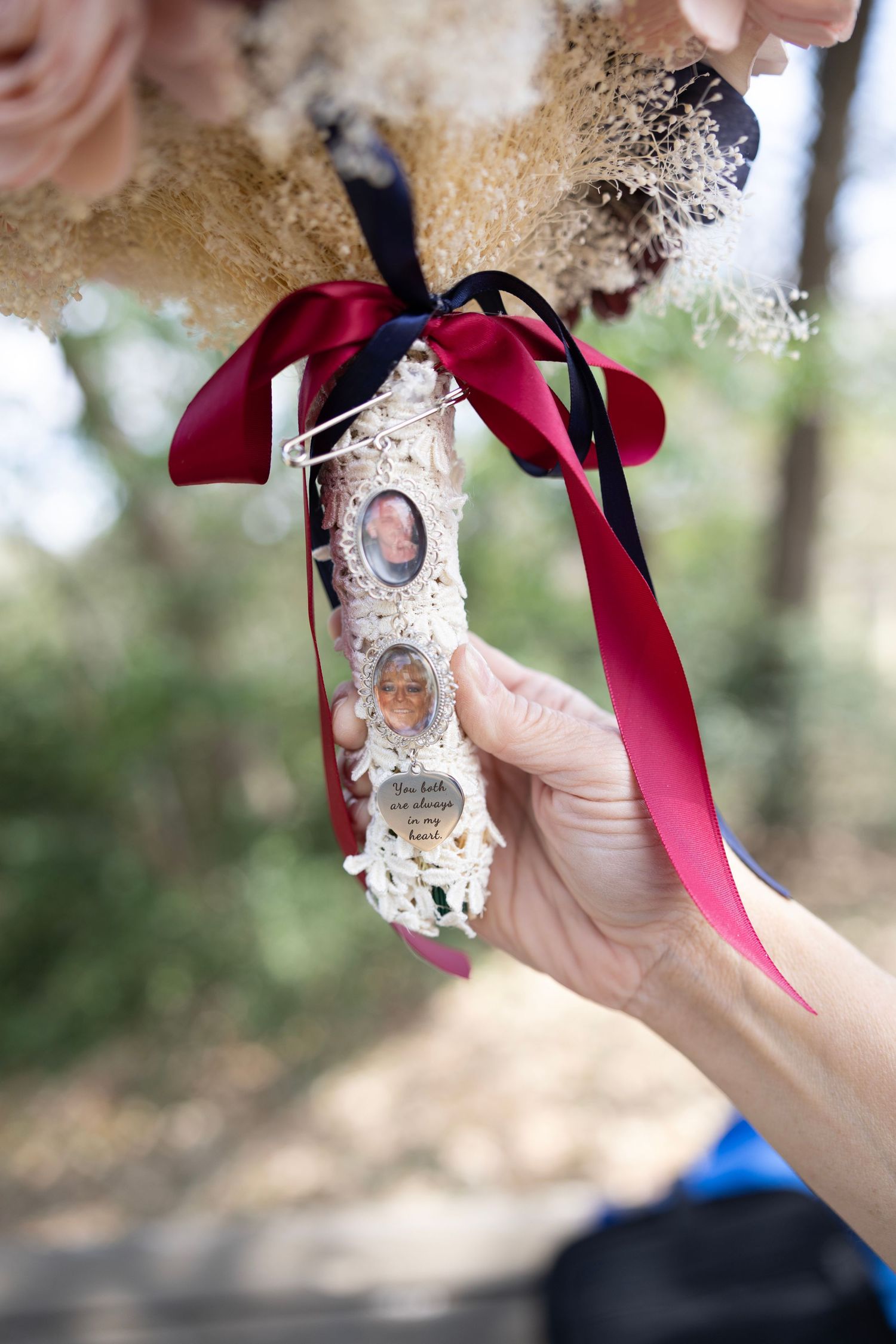 Hand holding a wedding bouquet charm with memorial photos and elegant red and black ribbons outdoors.