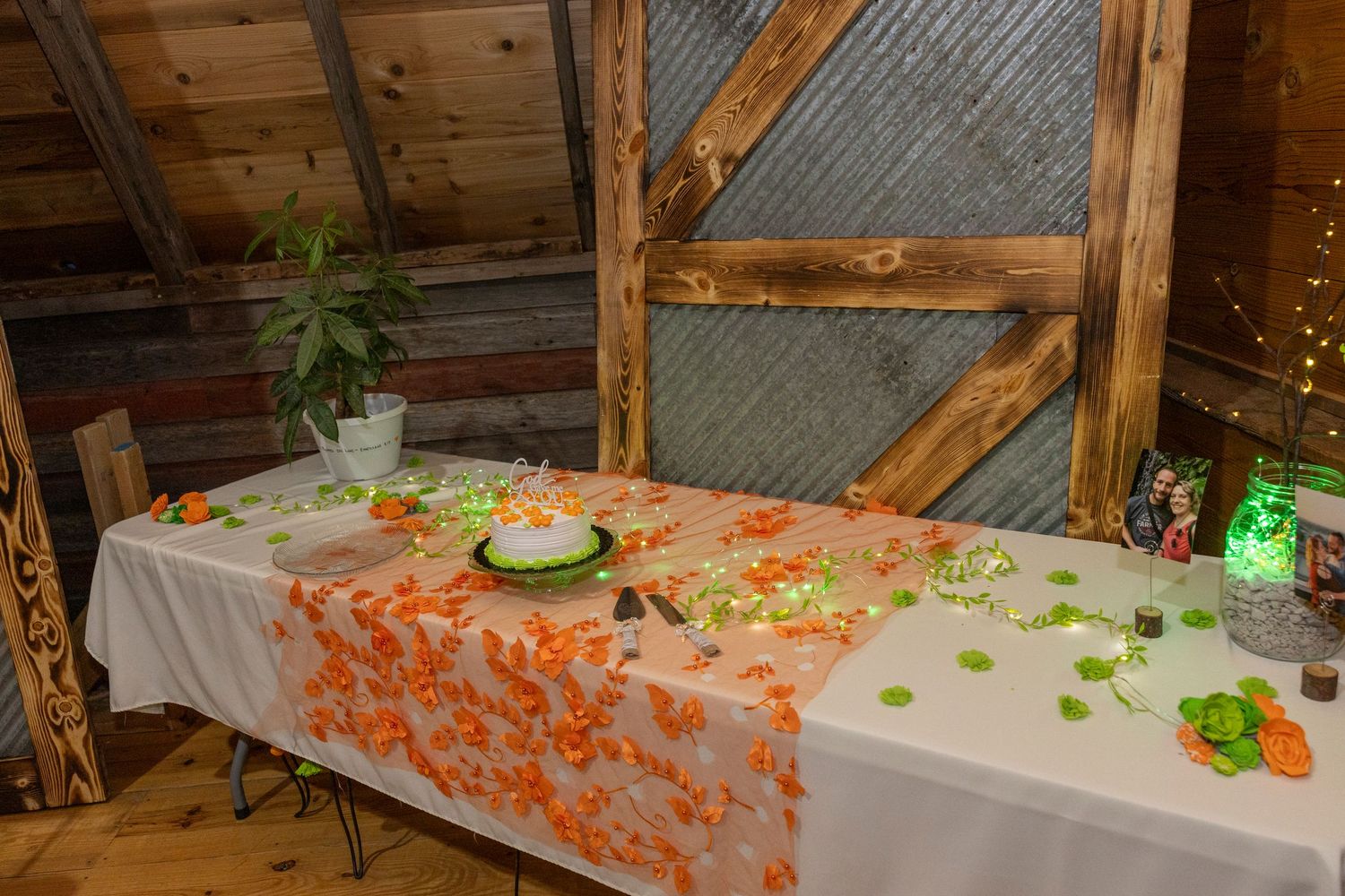Rustic barn reception table with orange floral runner, decorated cake, and festive party decor on white tablecloth.