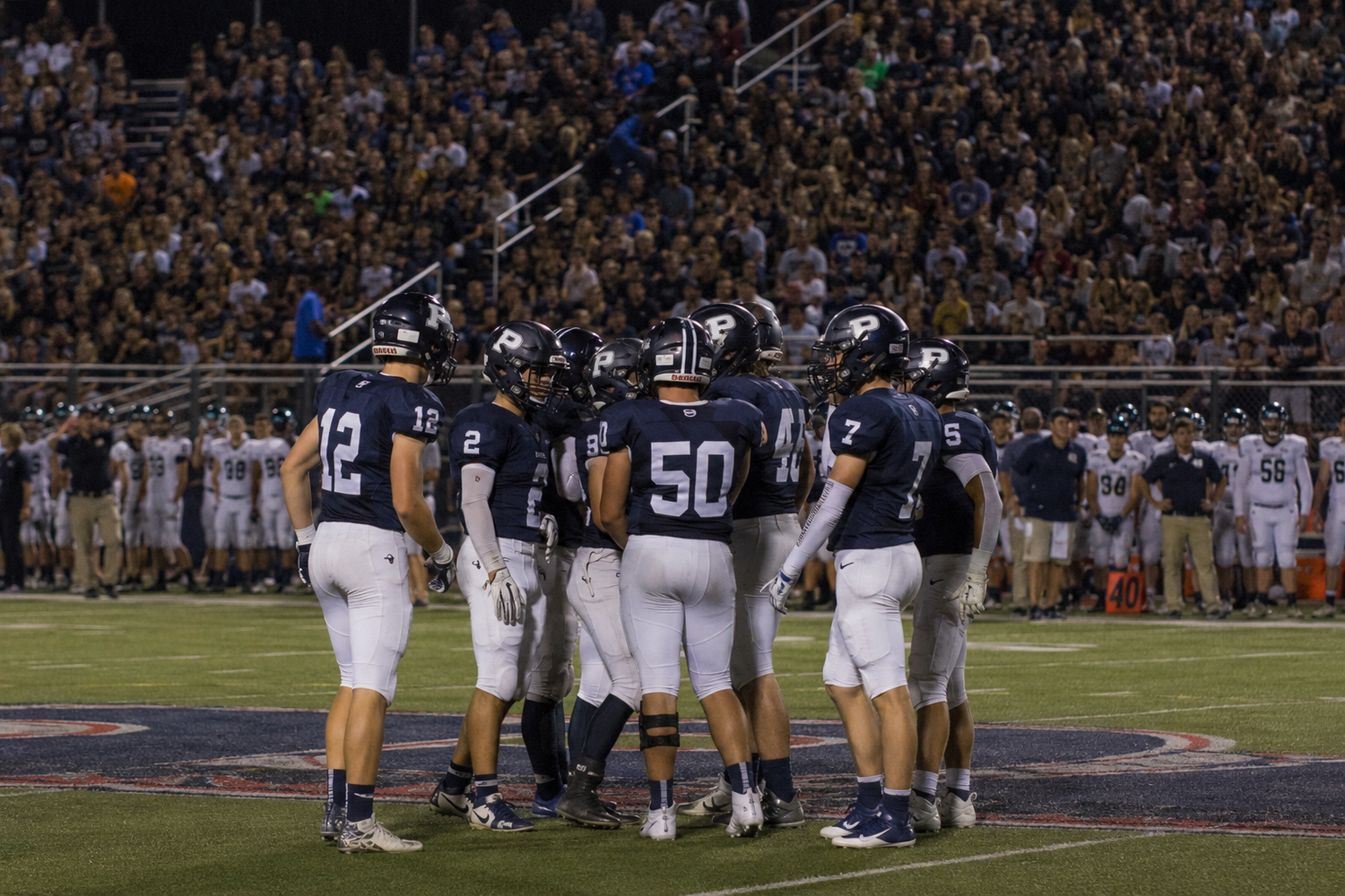 A high school football team huddles on the field during a night game with packed bleachers behind them.