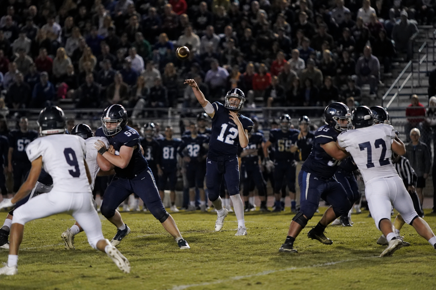 High school quarterback number 12 throws a pass during a night football game with blockers in formation.
