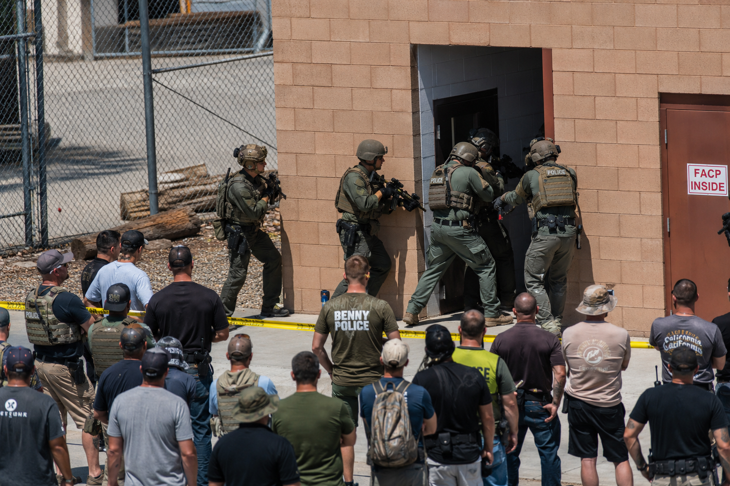 SWAT team officers in tactical gear conduct a building entry drill while observers watch behind yellow tape.