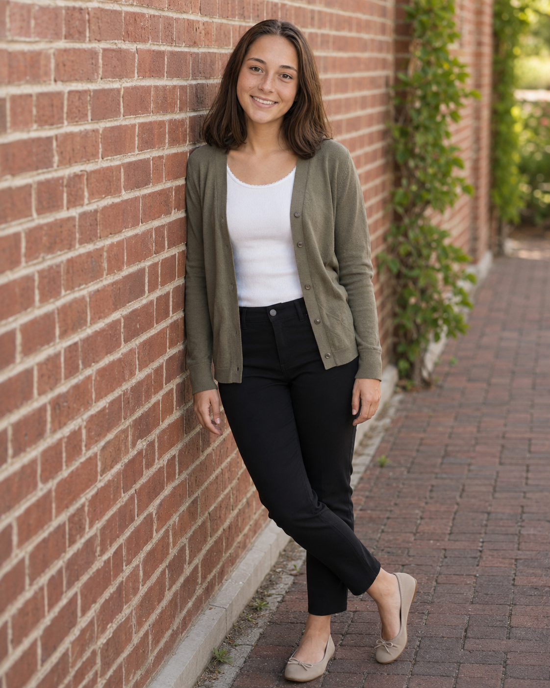 Young woman in olive cardigan and black jeans leaning against brick wall outdoors.