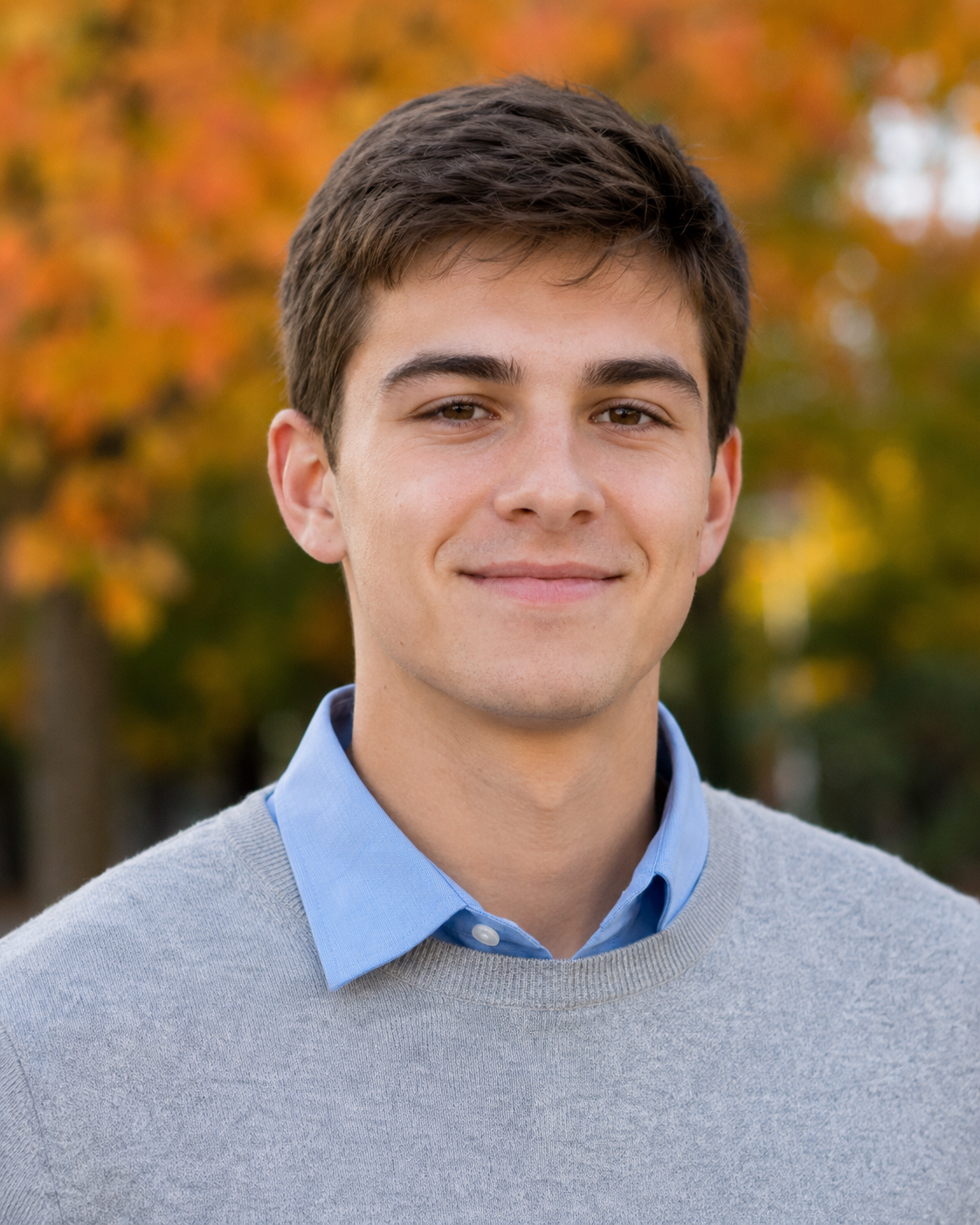 Smiling young man in gray sweater and blue collar shirt with fall foliage background.