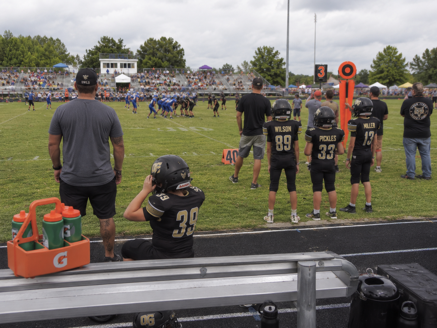High school football players and coaches watch from the sideline during a sunny afternoon game.