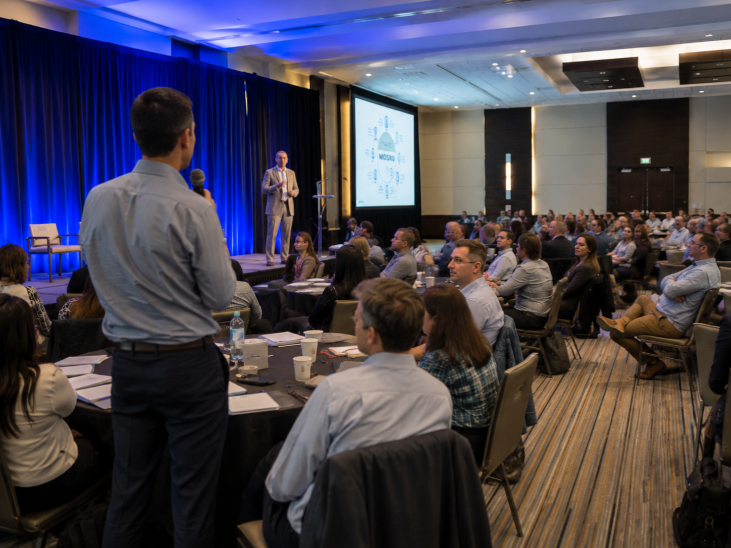 A speaker presents to a large audience at a professional conference in a hotel ballroom with blue lighting.