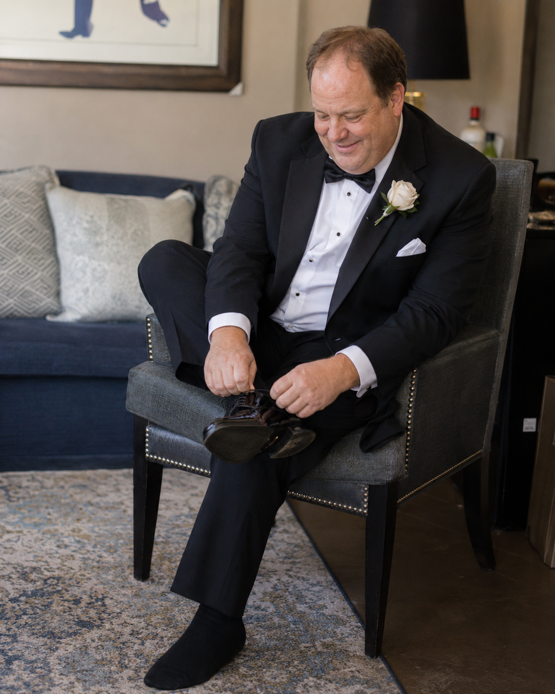 Dapper groom in black tuxedo with white boutonniere puts on dress shoes while seated in armchair.