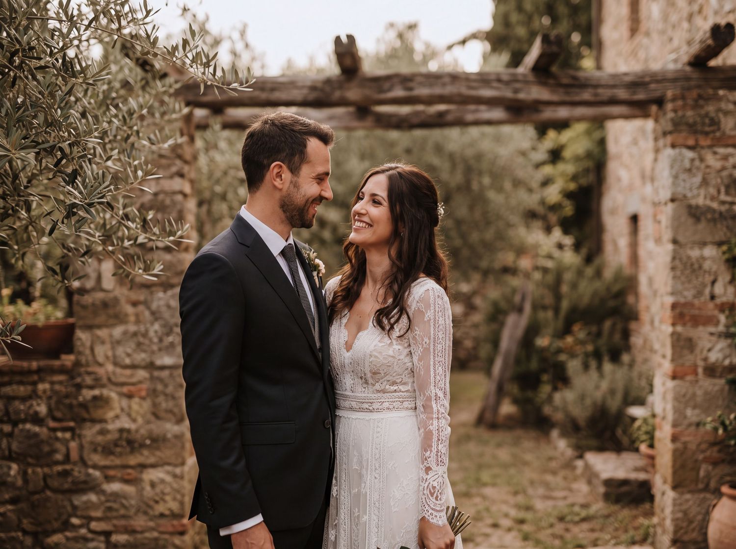 A smiling bride and groom embrace outdoors at a rustic stone villa wedding venue surrounded by olive trees.