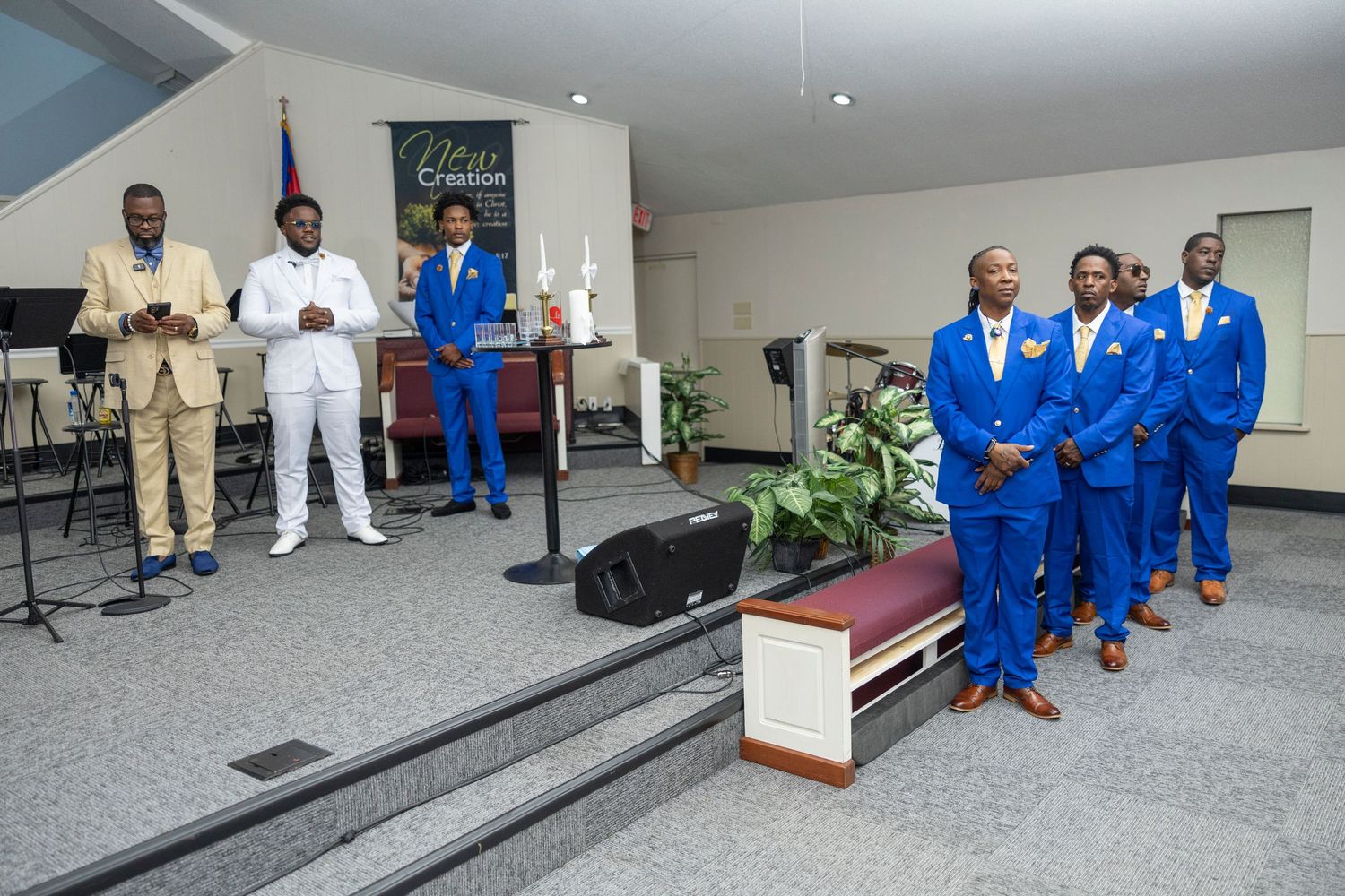 Men in blue suits stand in a church during a formal ceremony, with two speakers at a podium near the altar.