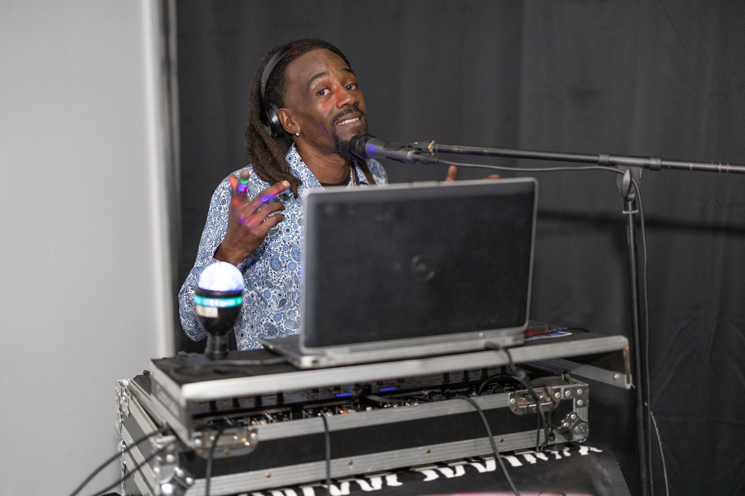 A DJ performing at an event, smiling while using a laptop and mixer equipment with a microphone stand nearby.