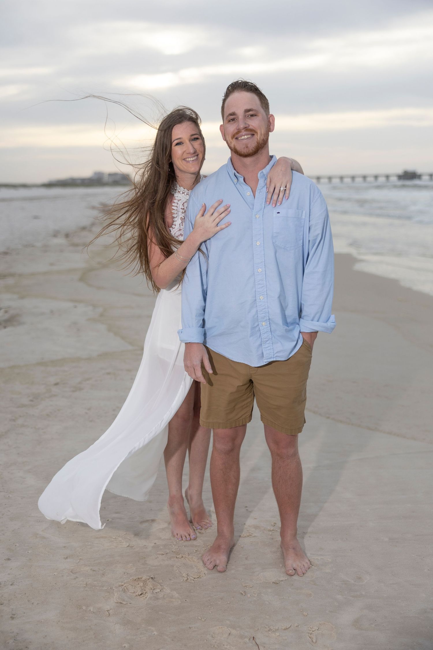 Couple posing barefoot on a sandy beach at sunset, woman in white dress and man in blue shirt and khaki shorts.