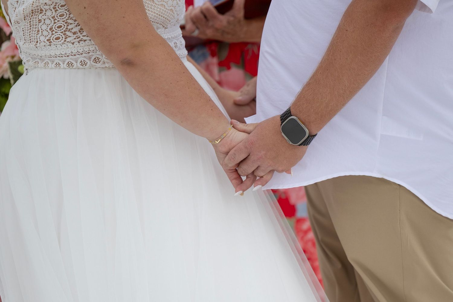 Bride and groom holding hands at wedding ceremony, bride in white lace gown, groom in white shirt with smartwatch.