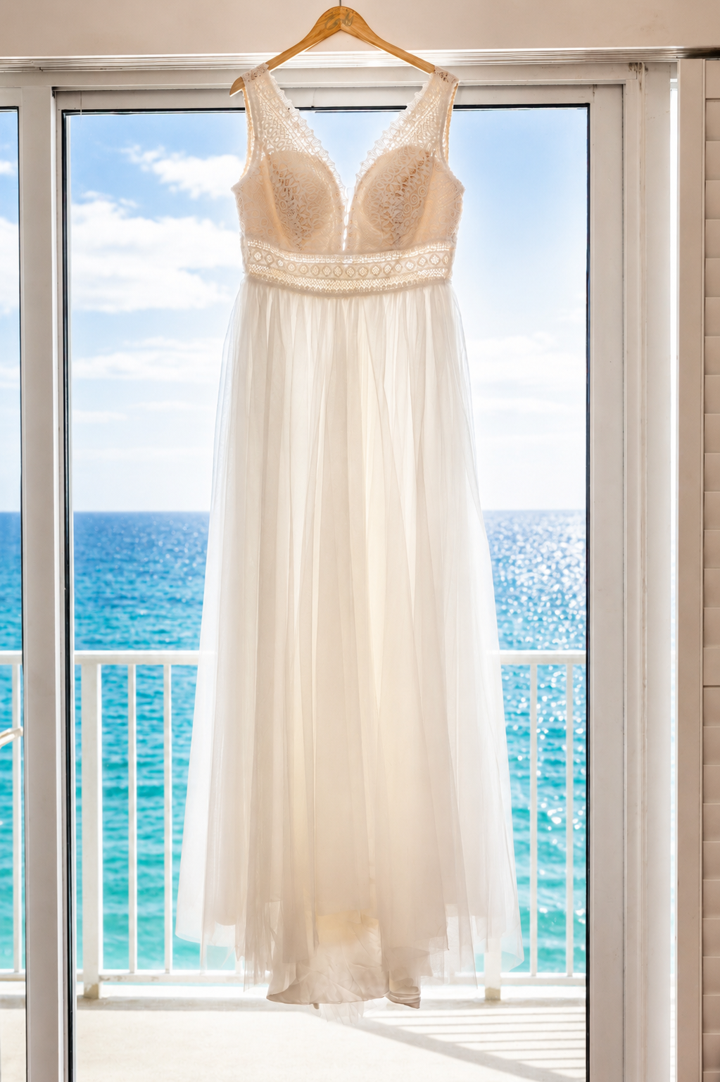 White lace wedding dress hanging on a wooden hanger in front of a balcony with turquoise ocean views.