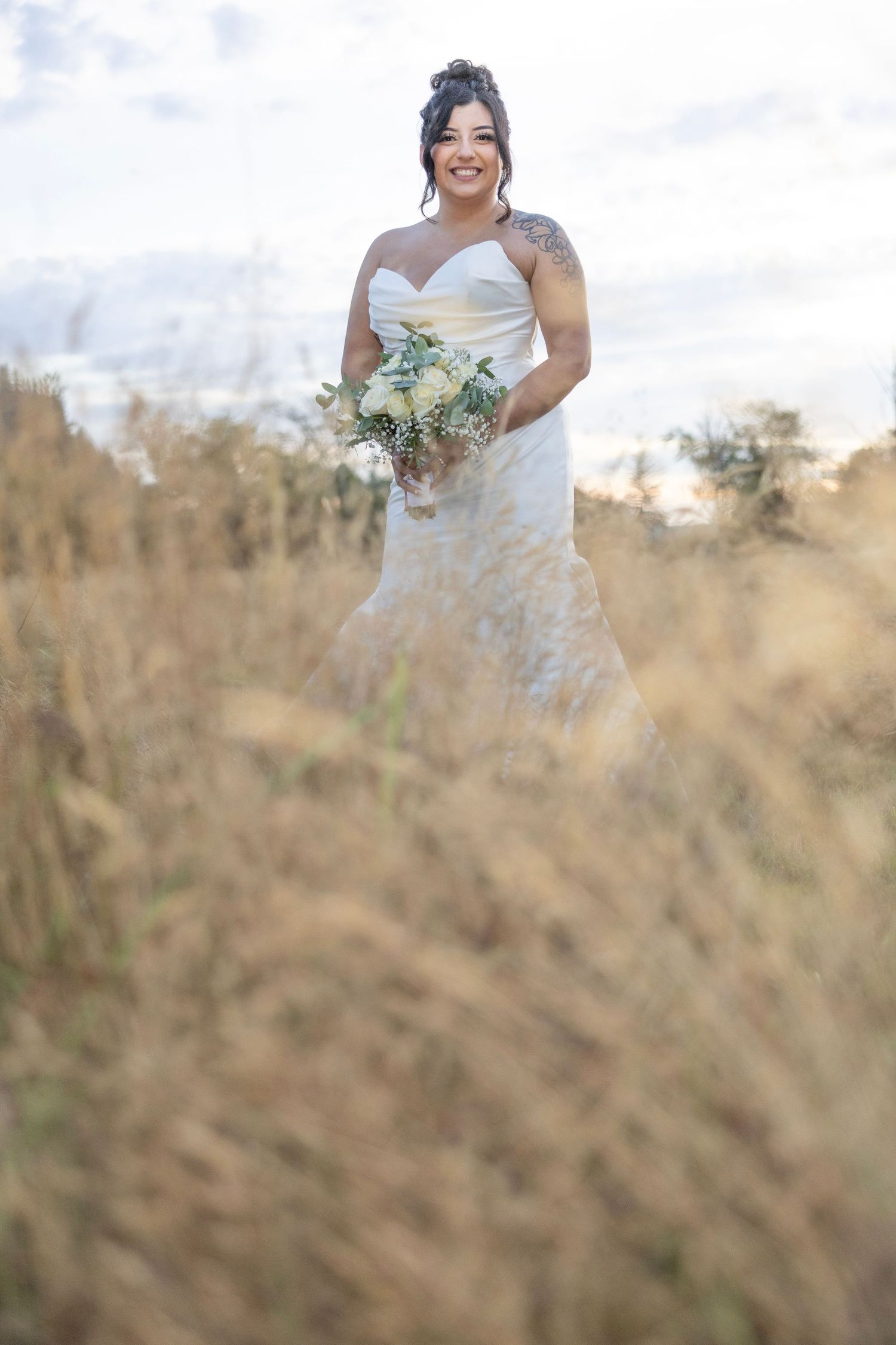 A bride in a white strapless wedding dress poses in a field of tall golden grass during sunset.