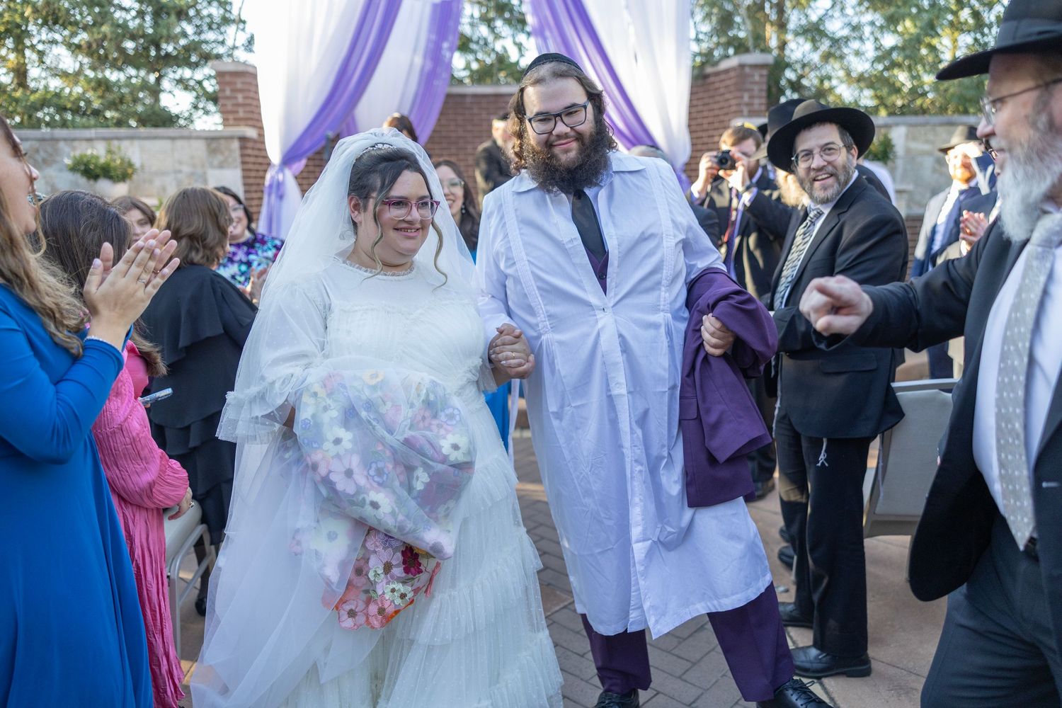 Traditional Jewish wedding ceremony taking place outdoors under a purple chuppah with guests celebrating.