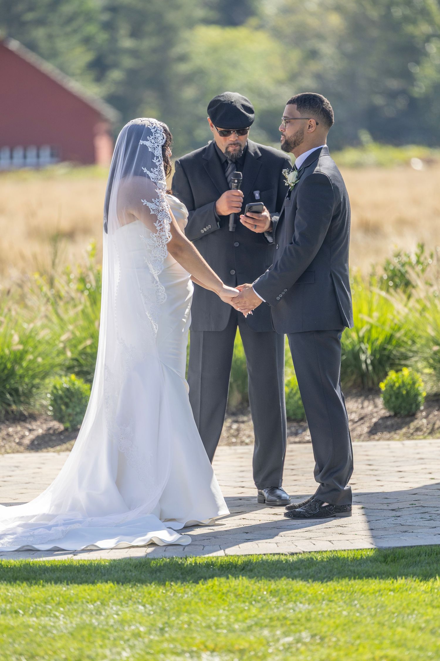 Wedding ceremony taking place outdoors on a sunny day with people gathered on a brick patio near grassy lawn.