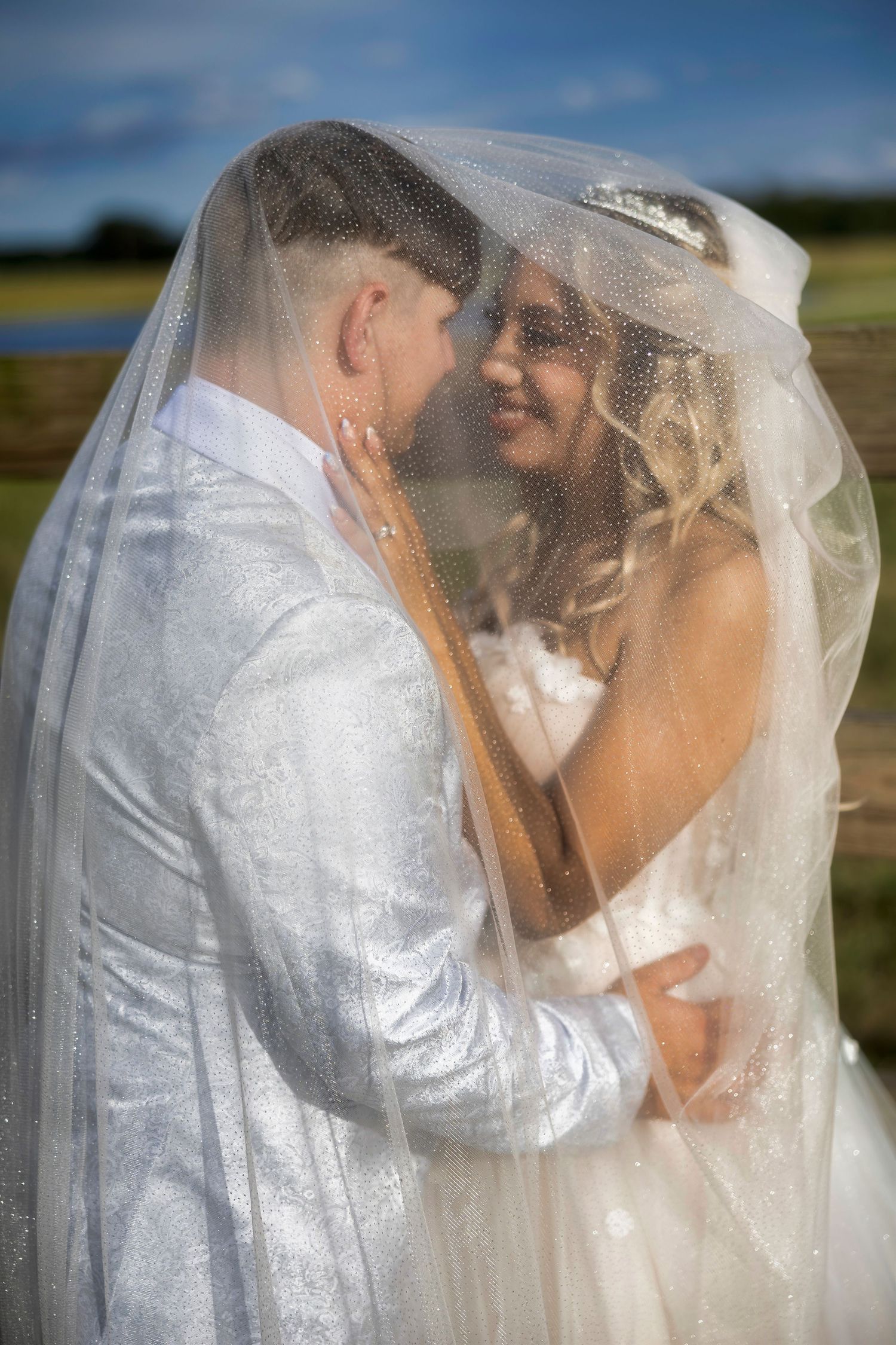 A romantic wedding moment captured through a flowing veil as newlyweds share an intimate embrace outdoors.