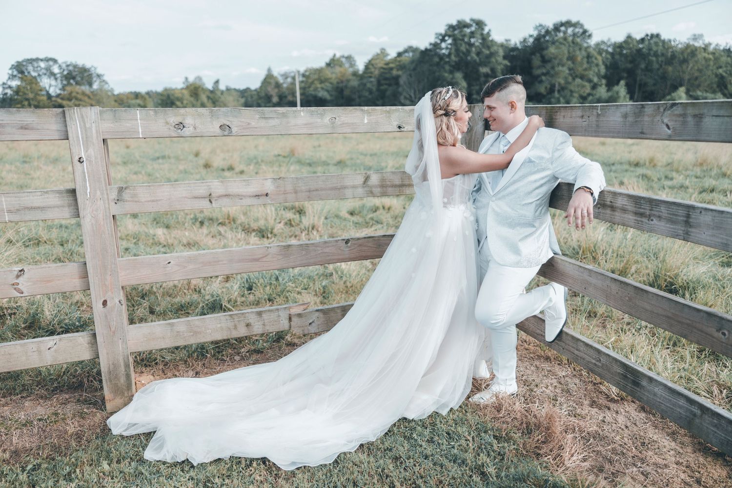 A couple in wedding attire share a romantic moment leaning against a rustic wooden fence in a countryside setting.