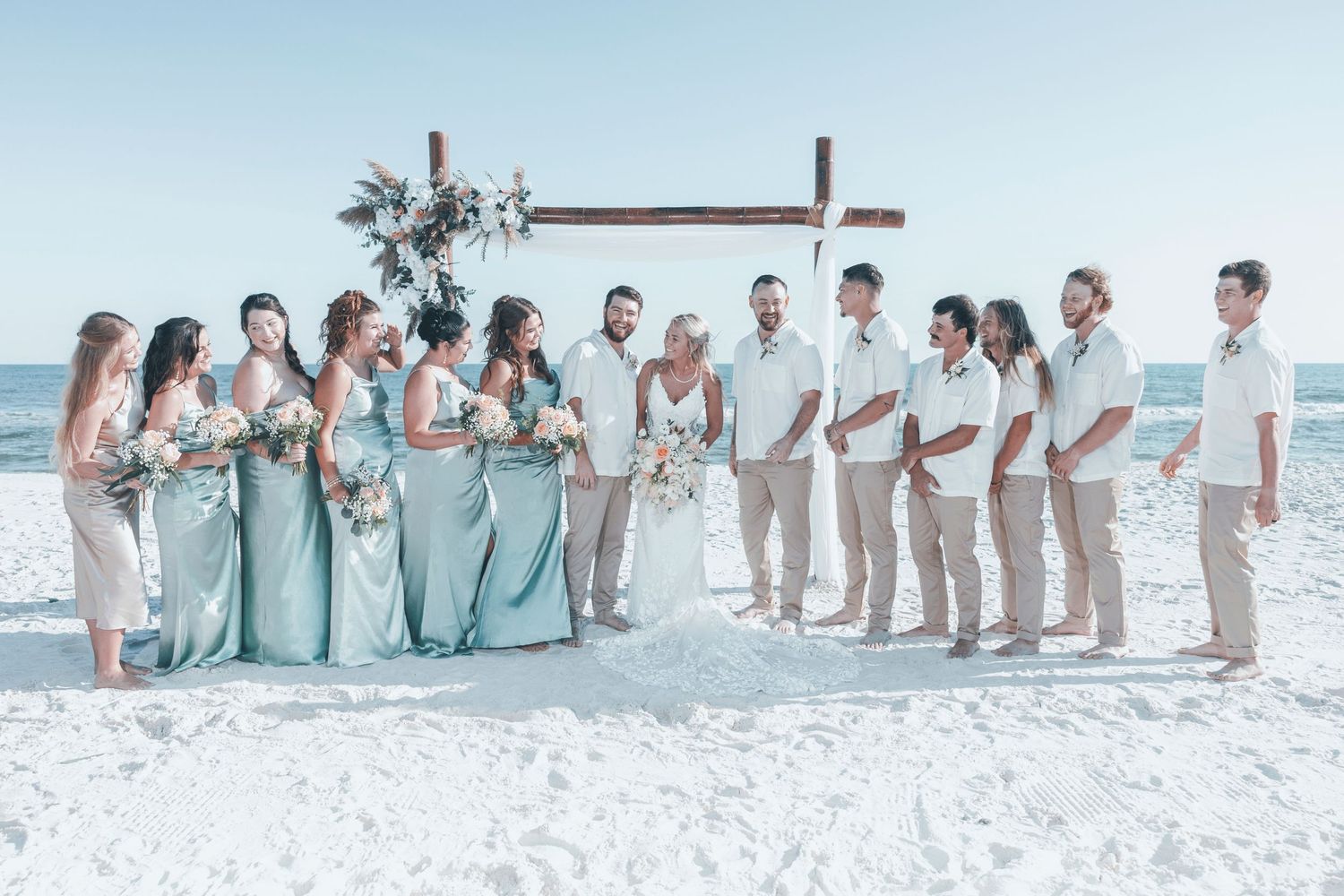 Wedding party in light blue and white attire poses together on a sunny beach with waves in the background.