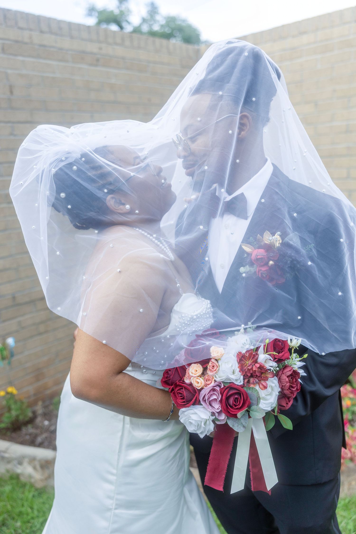 A romantic wedding couple embraces under a flowing white veil against a brick wall backdrop.