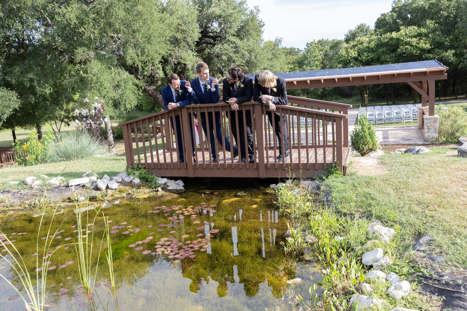 A group stands together on a wooden bridge over a tranquil pond surrounded by lush greenery and landscaping.