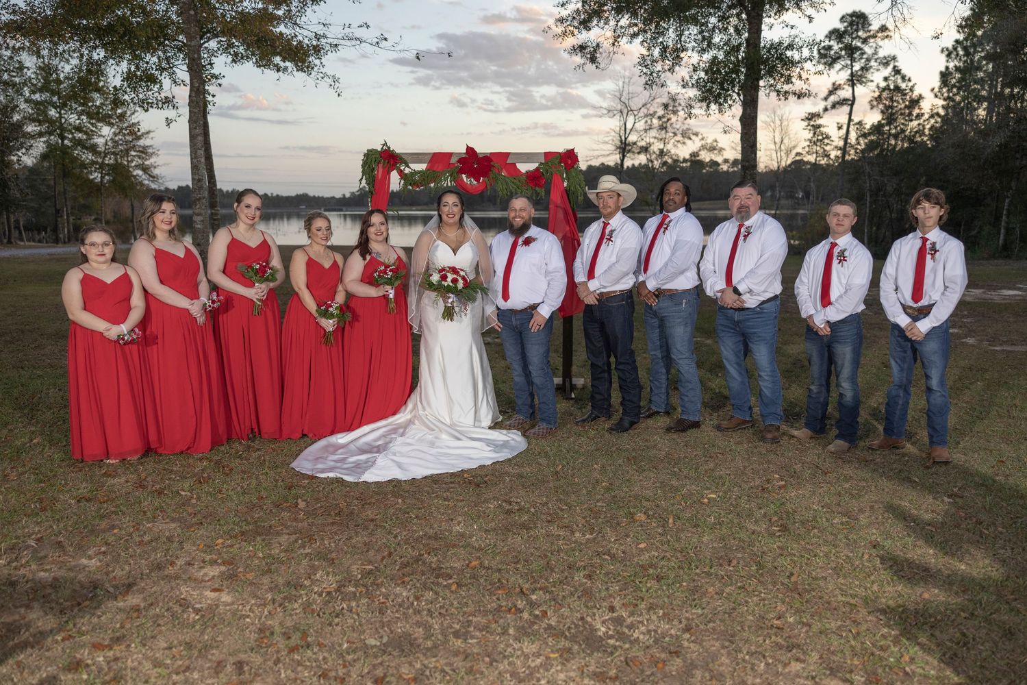 Wedding party in red dresses and blue jeans poses outdoors at sunset for rustic country-themed group photos.