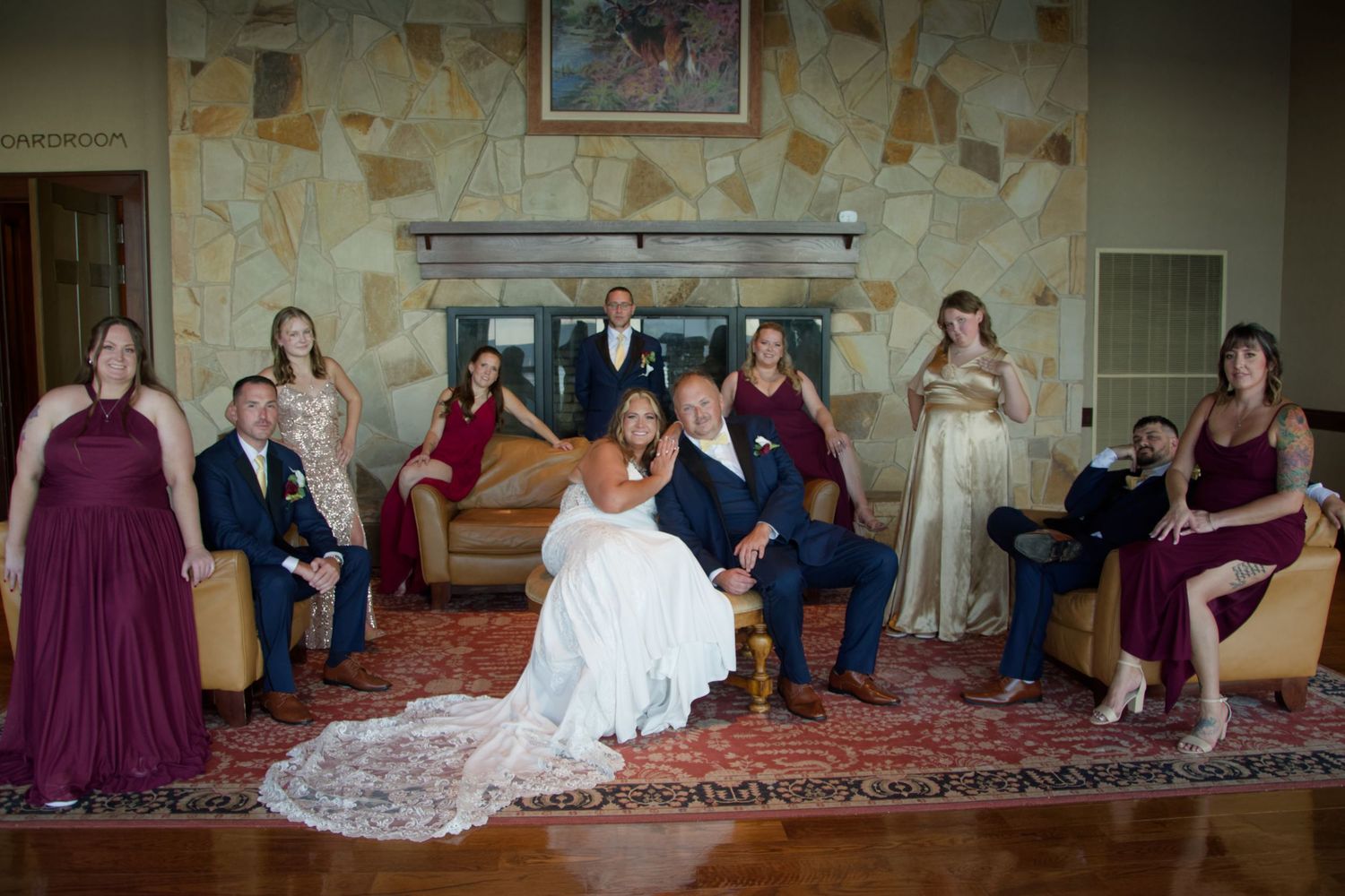 Wedding party poses playfully on floor in elegant venue with burgundy bridesmaids dresses and white bridal gown.