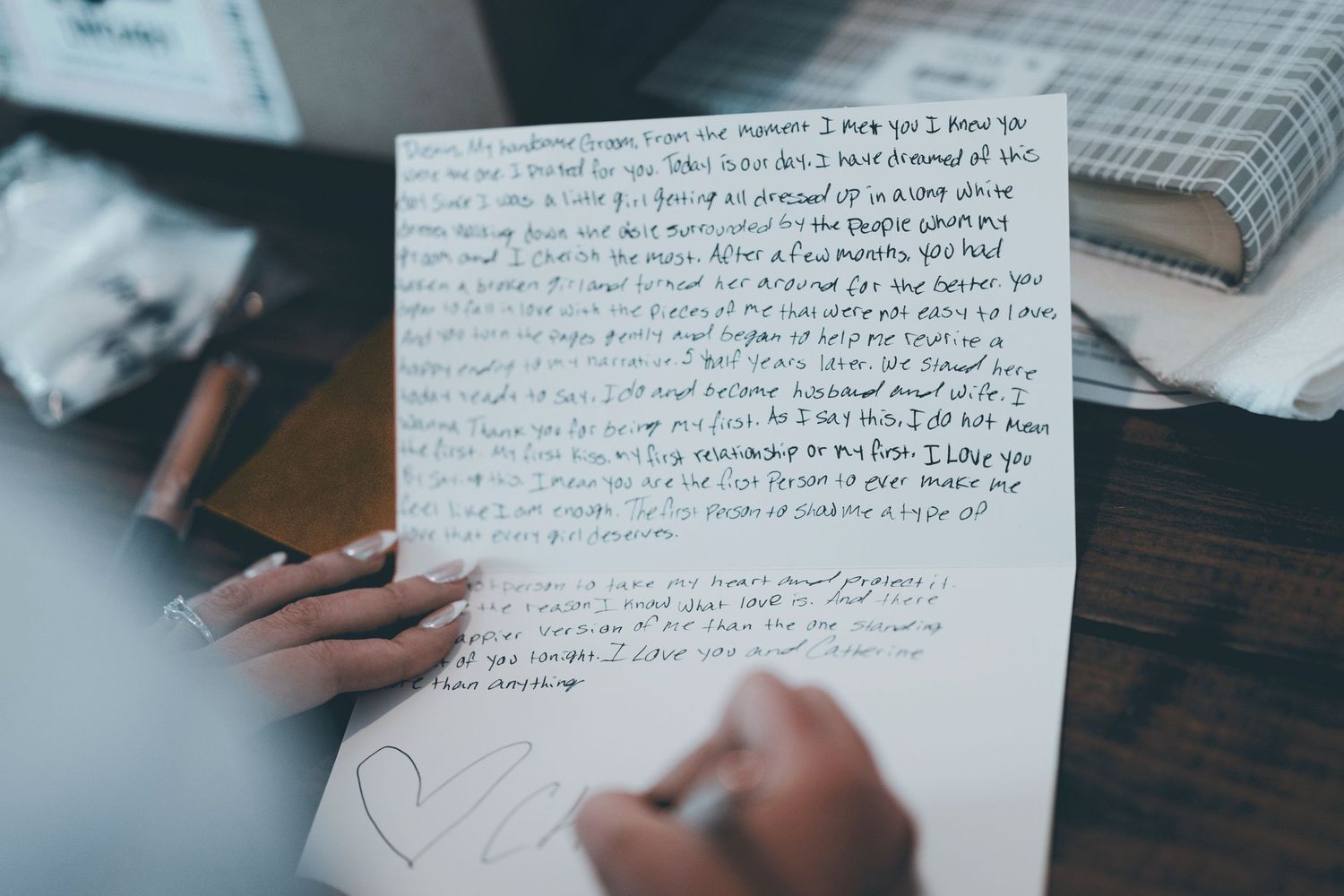 A person's hands writing on white paper with handwritten text visible, surrounded by notebooks and pencils on a desk.