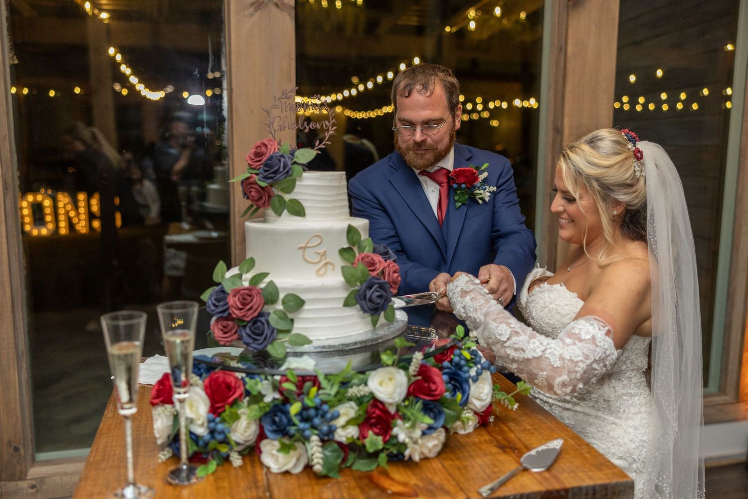 Newlyweds cut their wedding cake decorated with red and blue flowers under twinkling string lights at rustic venue.